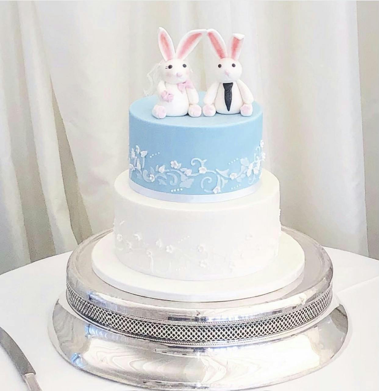 White and blue wedding cake with fondant bunny rabbits dressed up as a bride and groom on top