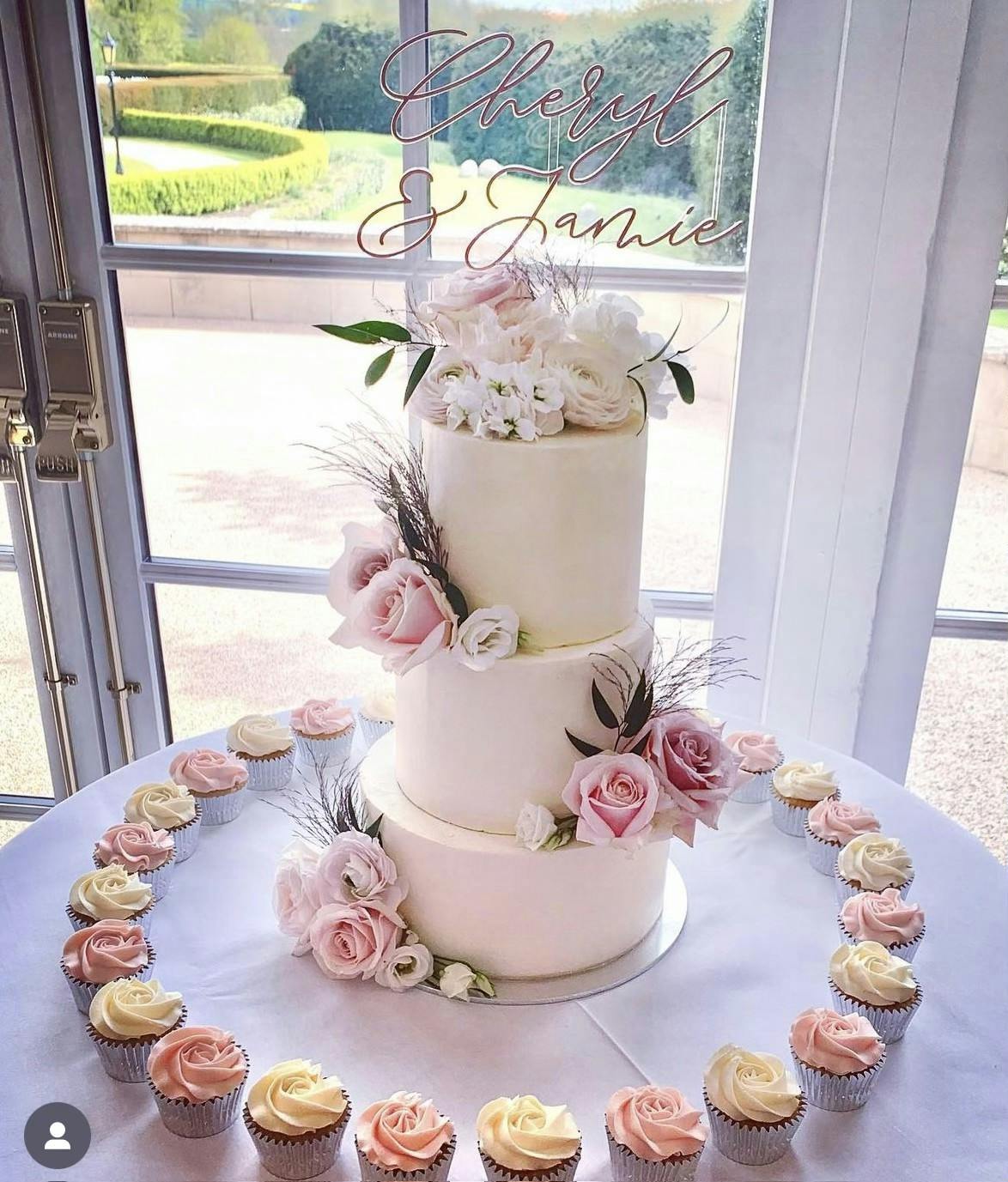 Light pink wedding cake with pink and white flowers surrounded by pink cupcakes for Cheryl and Jamie