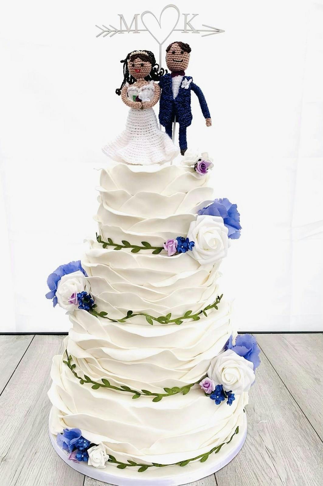 White wedding cake with white flowers, blue flowers, wavy icing patterns and knitted figures on top