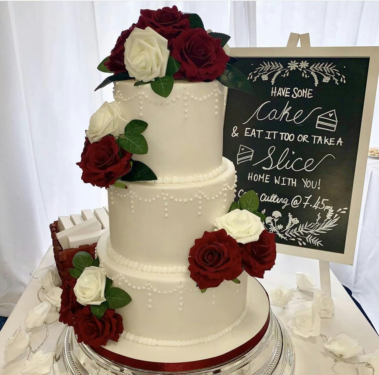 White wedding cake with red roses, white roses and cake pearls next to a chalk board